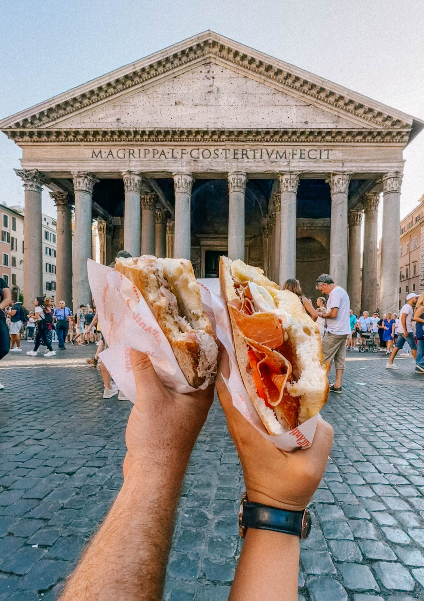 Two people hold up their Italian sandwiches at All'Antico Vinaio with the Pantheon in the background.