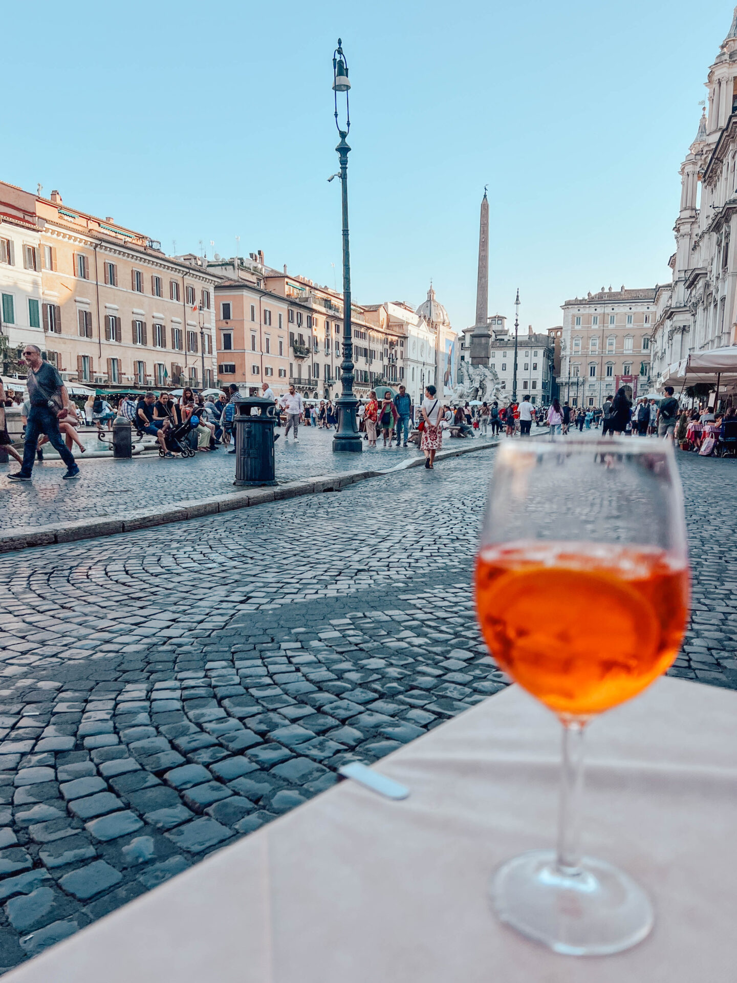 An aperol spritz sits on a table with the bustling Piazza Navona in the background