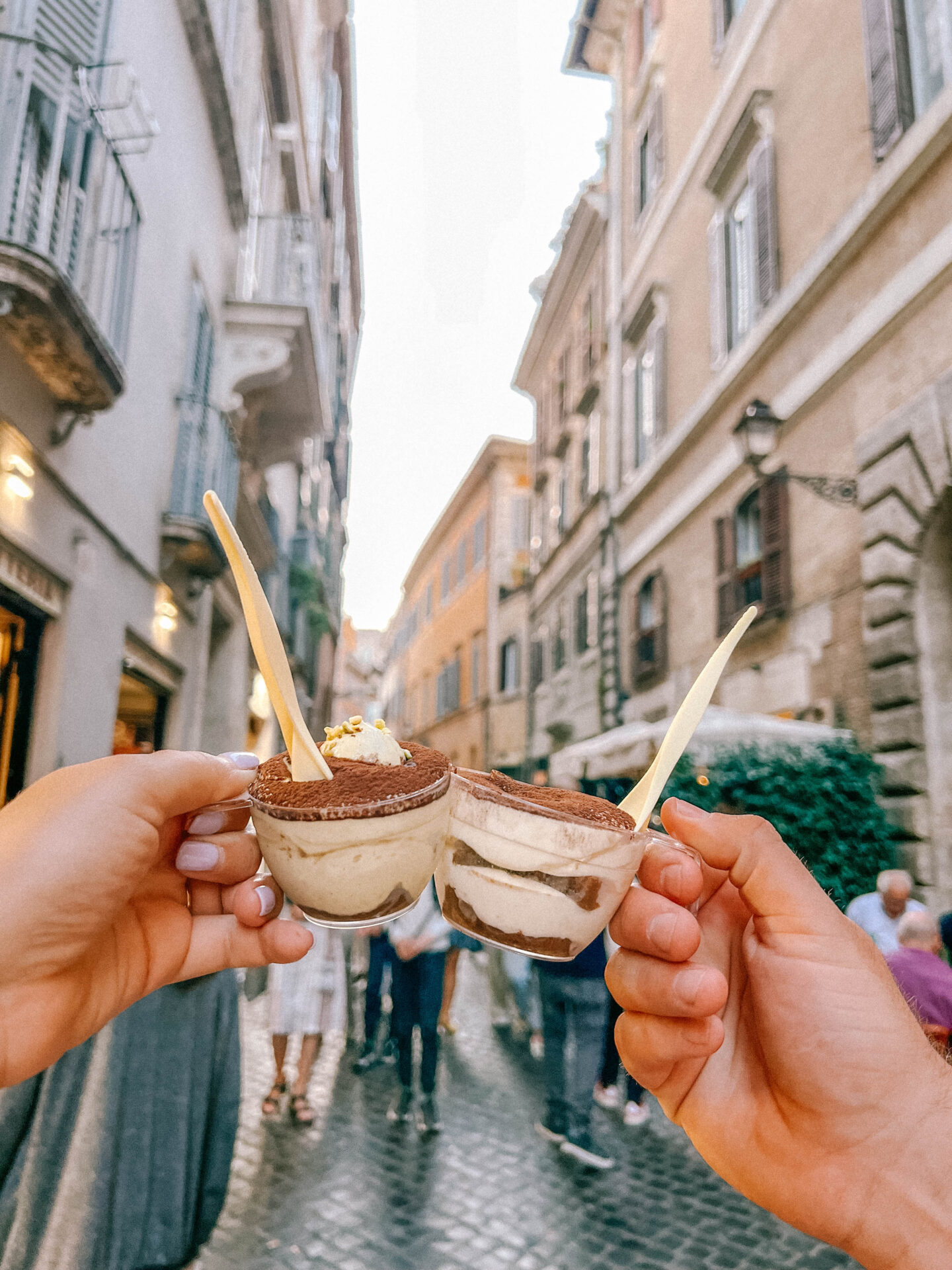 Two hands hold up cups of take-away tiramisu with the cobble-streets of Rome in the background