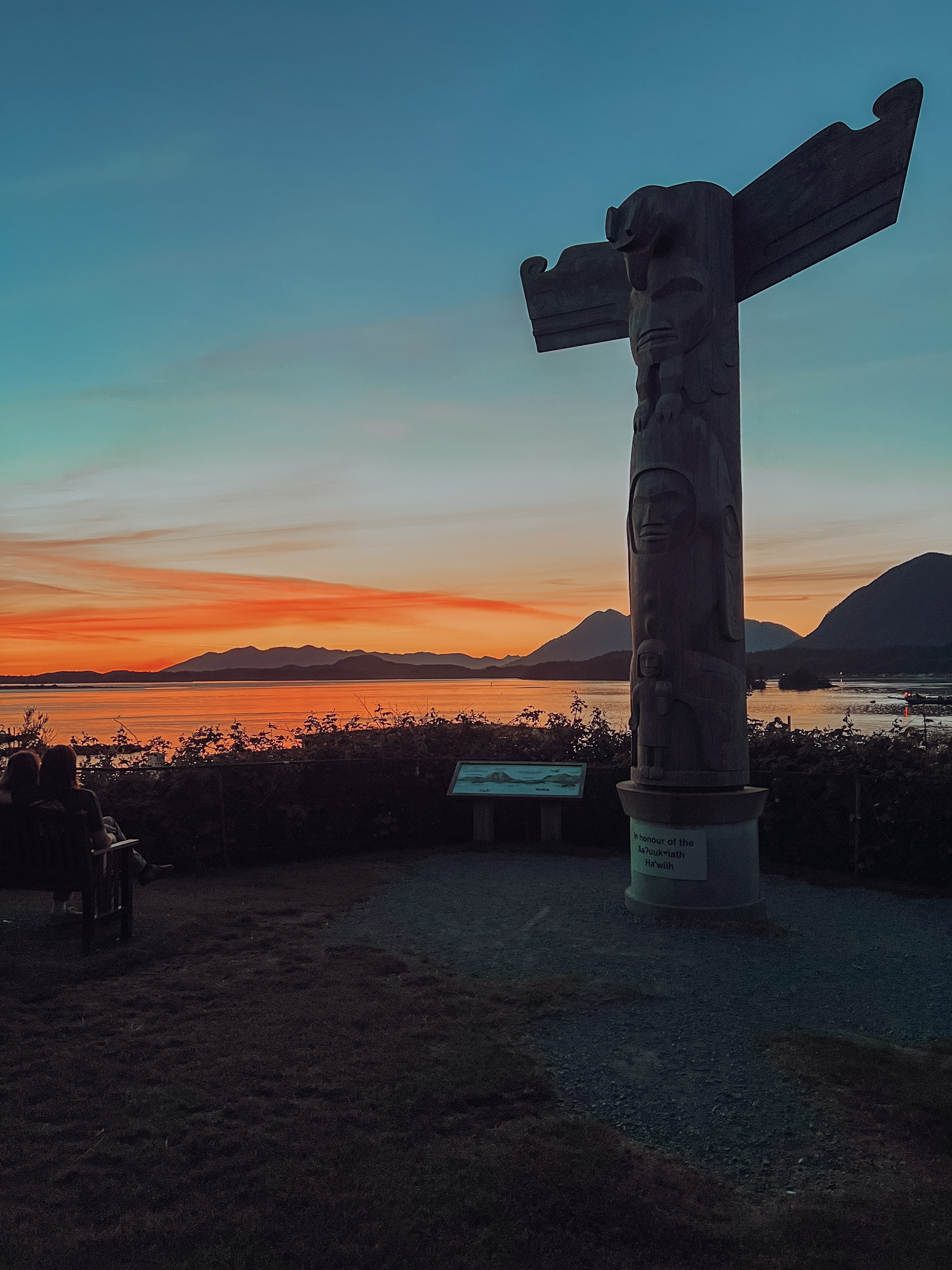 An indigenous totem pole stands in front of a beautiful orange sunset view of Tofino's inlet.