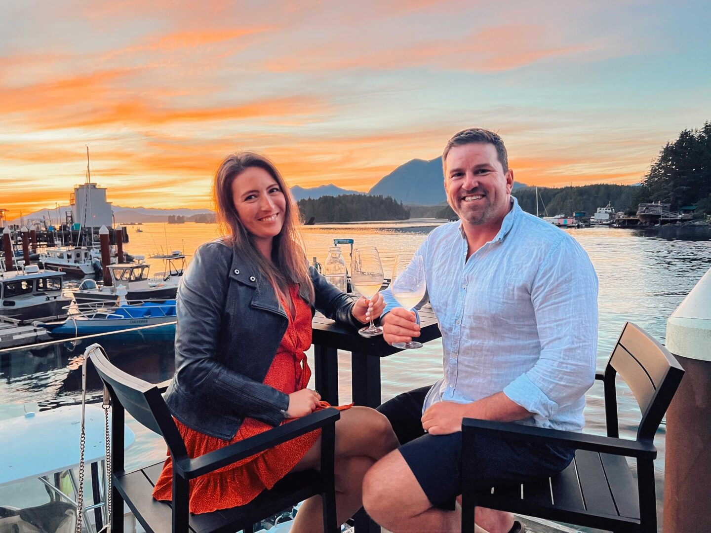 A happy couple cheers on Shelter's beautiful waterfront Restaurant in Tofino with boats and the sun setting in the background.