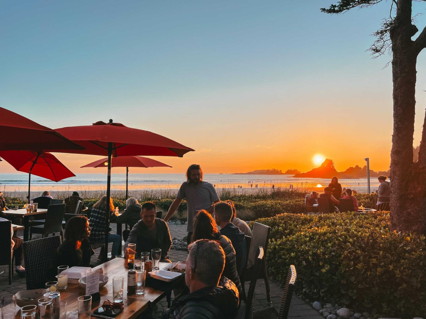 Friends gather around a table at the Sandbar Bistro at Long Beach Lodge, while the sun sets over Cox Bay Beach.