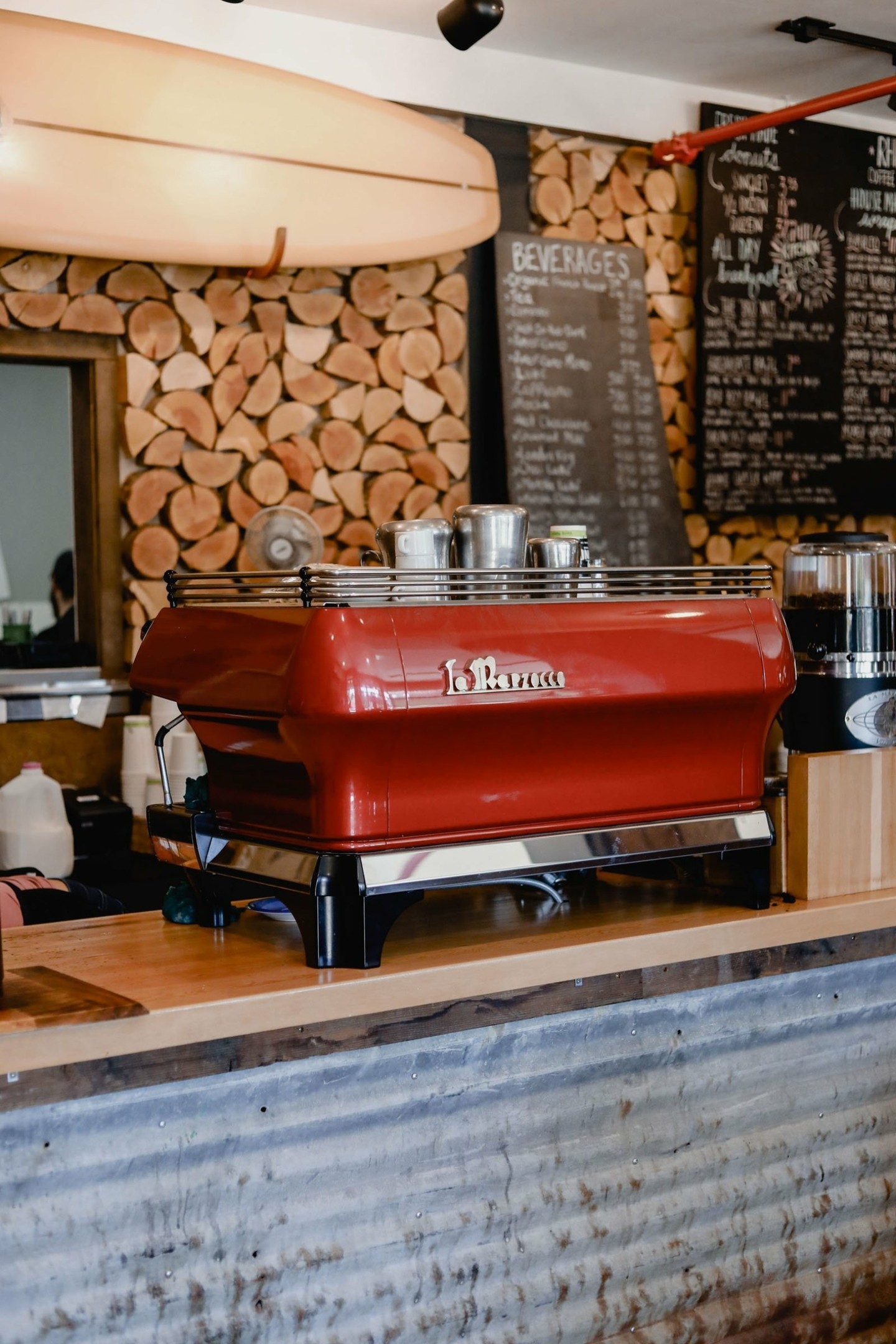 A beautiful Italian red espresso machine sits on a bar at Rhino's Coffee House. In the background there is a surfboard menus and wood slabs on the wall. 