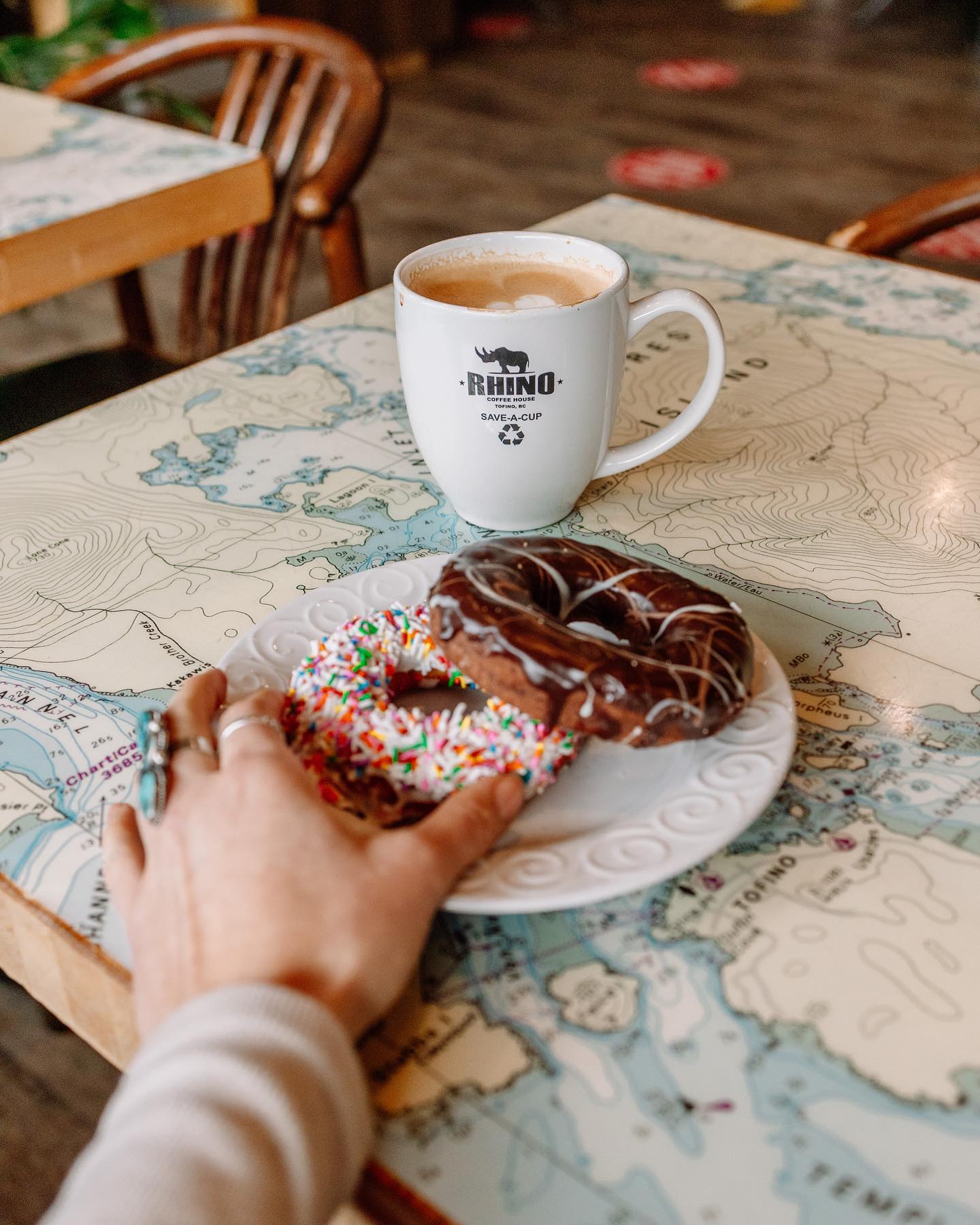 A girl with rings on reaches out to two donuts on a plate and a mug of coffee that says Rhino's coffee House on it. Everything is rest on a table with a map on it. 